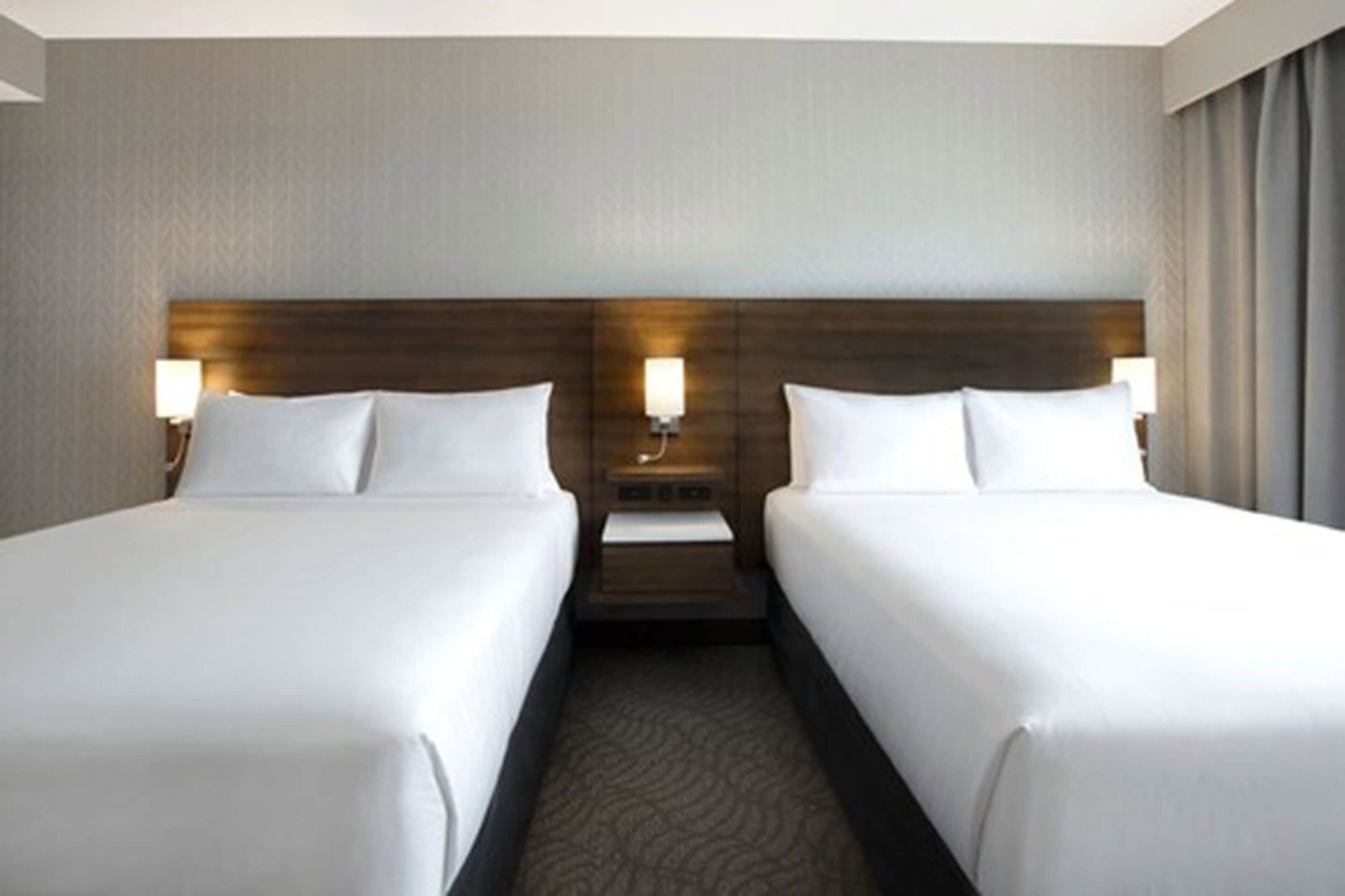 A hotel room with two beds, a wooden headboard, a nightstand with a lamp, and a carpeted floor.
