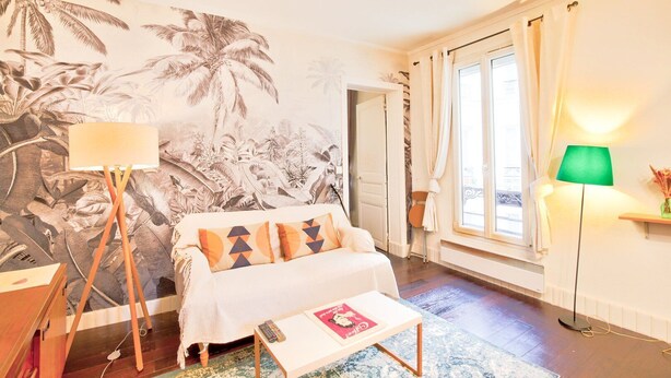 Een kamer met een grote tropische muurschildering, een witte bank met gestreepte kussens, een klein tafeltje met een afstandsbediening en een groene vloerlamp.