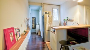 Een smalle keuken met een houten aanrecht, een koelkast en een spoelbak. Er hangen lampen boven het aanrecht en een spiegel aan de muur.