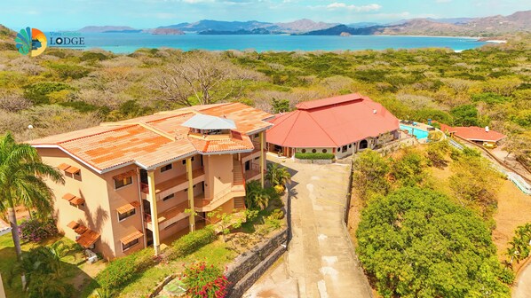 A multi-story building with a red roof, surrounded by lush greenery and a clear view of a lake and mountains in the distance. - Vista Condor Lodge (Cabo Velas)
