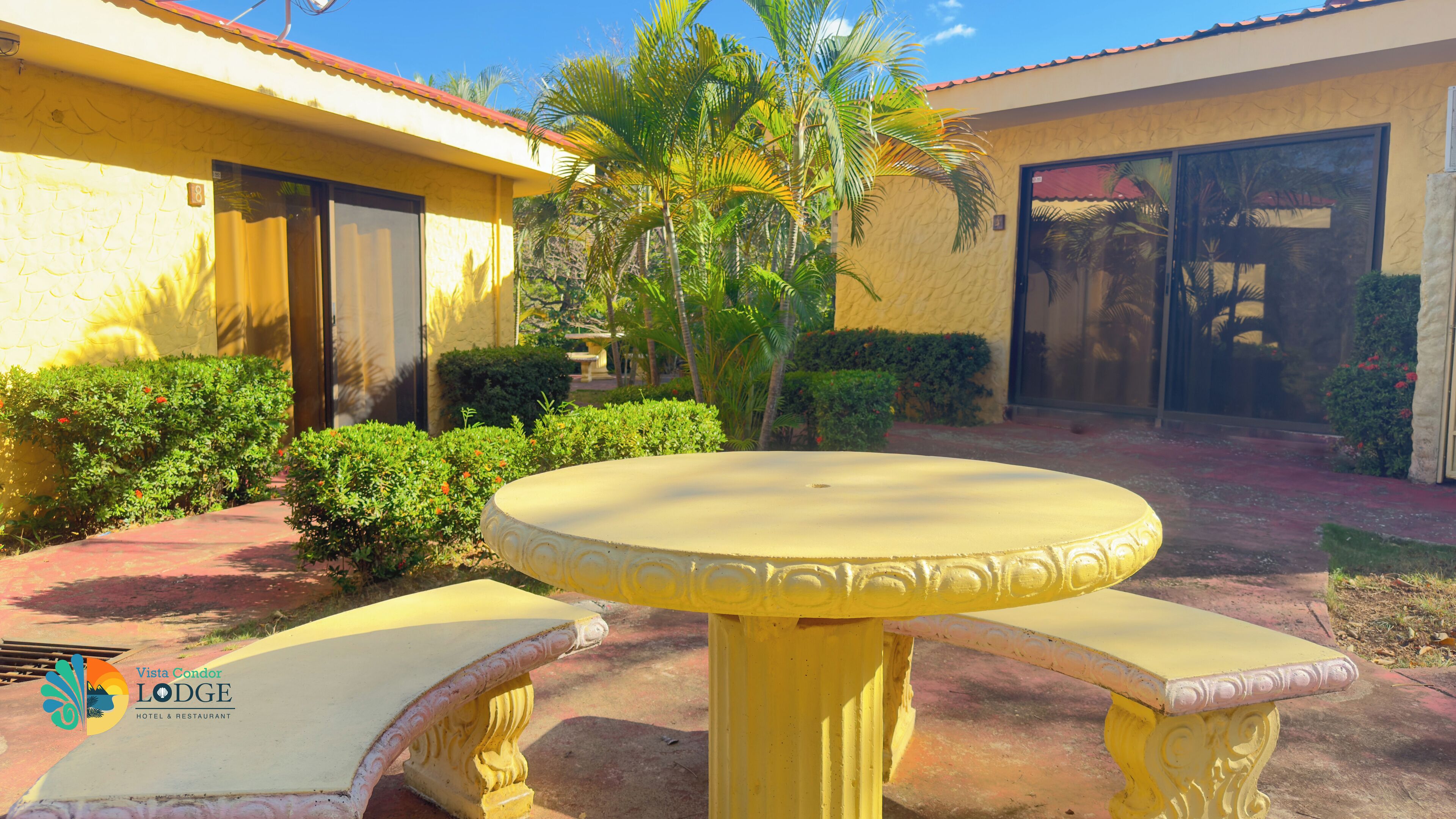 A yellow outdoor seating area with a round table and benches, surrounded by greenery and a building with a red roof.