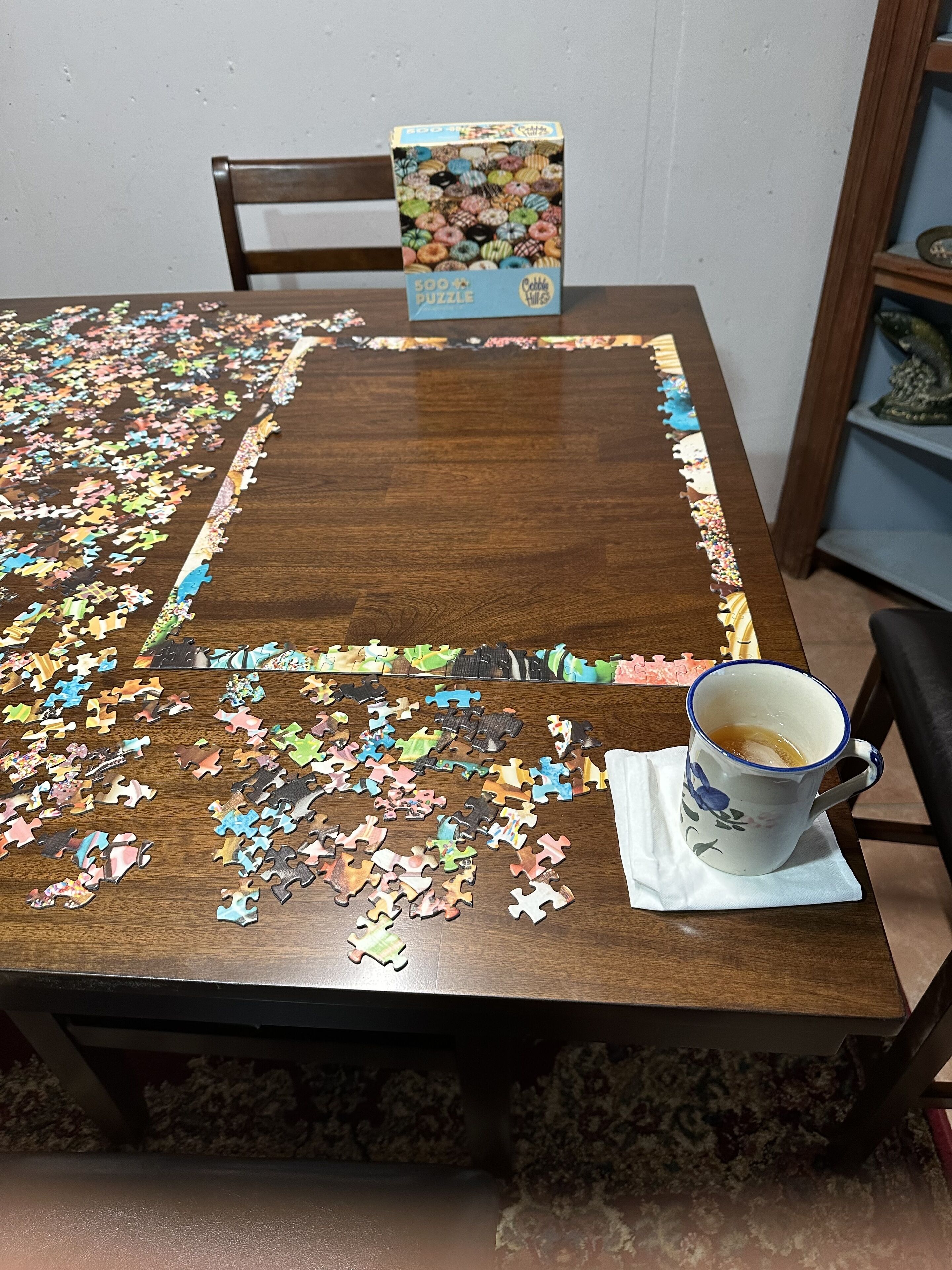 A wooden dining table with a 500-piece puzzle partially completed, a box of the puzzle, and a mug on a coaster.