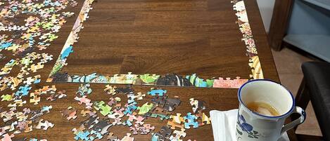 A wooden dining table with a 500-piece puzzle partially completed, a box of the puzzle, and a mug on a coaster.