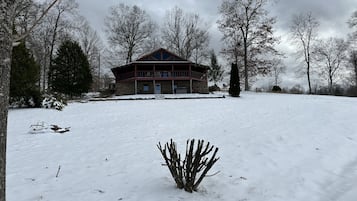 Eine große, zweistöckige Holzhütte mit Steinunterbau und rotem Dach, umgeben von verschneitem Gelände und kahl stehenden Bäumen.