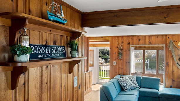 A cozy room with wooden walls, a blue sofa, and a sign reading "BONNET SHORES 41.4736° N, 71.4229° W".