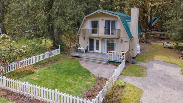 Une cabane à deux étages dotée d’un balcon, entourée d’une clôture en bois peinte en blanc, située dans une zone boisée.