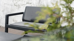 A black metal bench with a mesh backrest, positioned against a white textured wall.