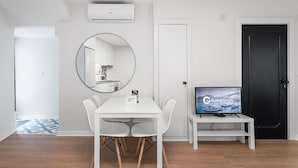 A modern, minimalist room with a white dining table, chairs, a round mirror, a flat-screen TV, and a black door.
