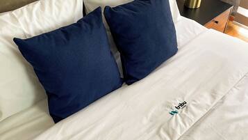 Una cama bien hecha con almohadas blancas y azul oscuro, un logotipo visible en la colcha y una mesita de noche con una lámpara y un objeto pequeño.