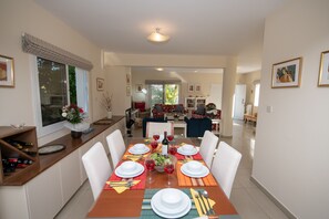 A dining area with a wooden table set for four, surrounded by white chairs. There is a kitchen area in the background with a dining table and chairs.
