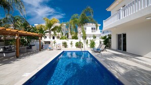 A rectangular swimming pool with clear blue water, surrounded by a white building with balconies, and a pergola with outdoor seating. - Graceful 3 Bedroom Agia Napa Villa with Pool (Agia Napa)