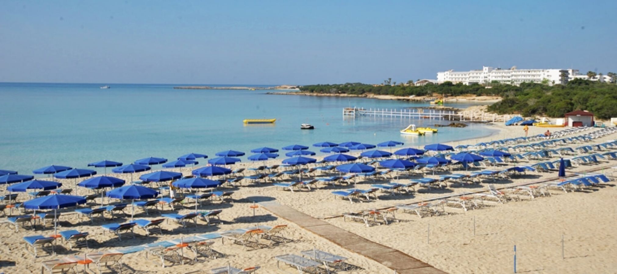 Une plage parsemée de nombreux parasols bleus et de chaises longues, une jetée qui s’avance dans l’eau et un bâtiment visible au loin.