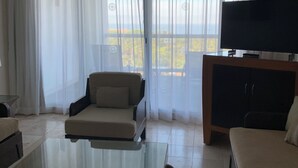 Un salon moderne avec une table basse en verre, un canapé, un téléviseur posé sur un meuble en bois et de grandes fenêtres ornées de rideaux légers.