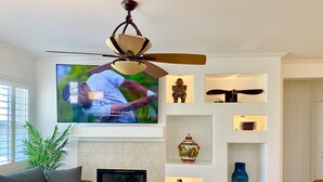 A modern living room with a flat-screen TV, a fireplace, decorative shelves, and a ceiling fan.