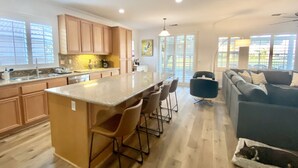 A modern kitchen with a central island, bar stools, and a dining area. There is a sofa and a pet bed with a dog in the living area.