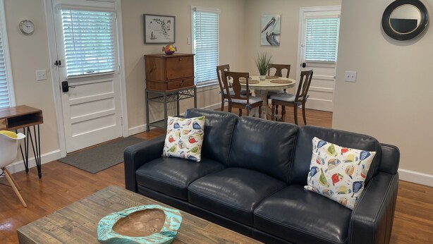 A living room with a black leather sofa, wooden coffee table, and a dining area with a round table and chairs. - Enjoy Crawfordville at the Courtyard’s Pelican Cottage. (Crawfordville)