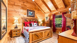 Quarto de cabana de madeira com cama, mesas de cabeceira, cômoda e janela com cortinas.