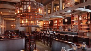 Interior de restaurante con vigas de madera, lámparas colgantes y una zona de bar con estantes llenos de botellas y vasos.