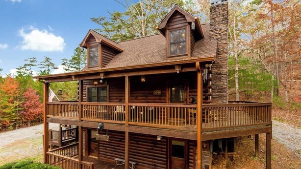 A two-story log cabin with a balcony, a stone chimney, and a porch area. - New Hot Tub Grt WIFI Pool Table Sleeps10 Family (Blue Ridge)