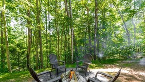 A forest setting with a fire pit and four Adirondack chairs arranged around it.