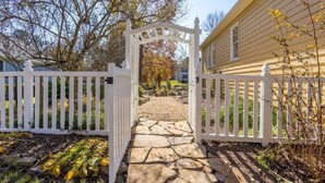A stone pathway leading to a white gate with a decorative arch, surrounded by a white picket fence and trees. - Hidden Gem Retreat | 2BR Basement |Near Mall of GA (Buford)