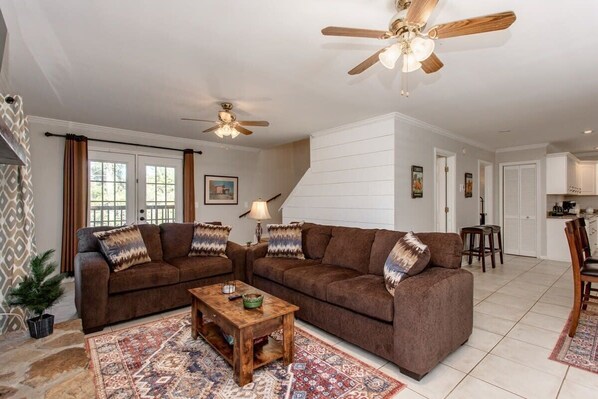 A living room with two brown sofas, a wooden coffee table, patterned rugs, ceiling fans, and a kitchen in the background. - Riverfront Home W/ Access| Fire Pit & Fire Place | Fiber Wifi| Slps 8 (Sautee Nacoochee)