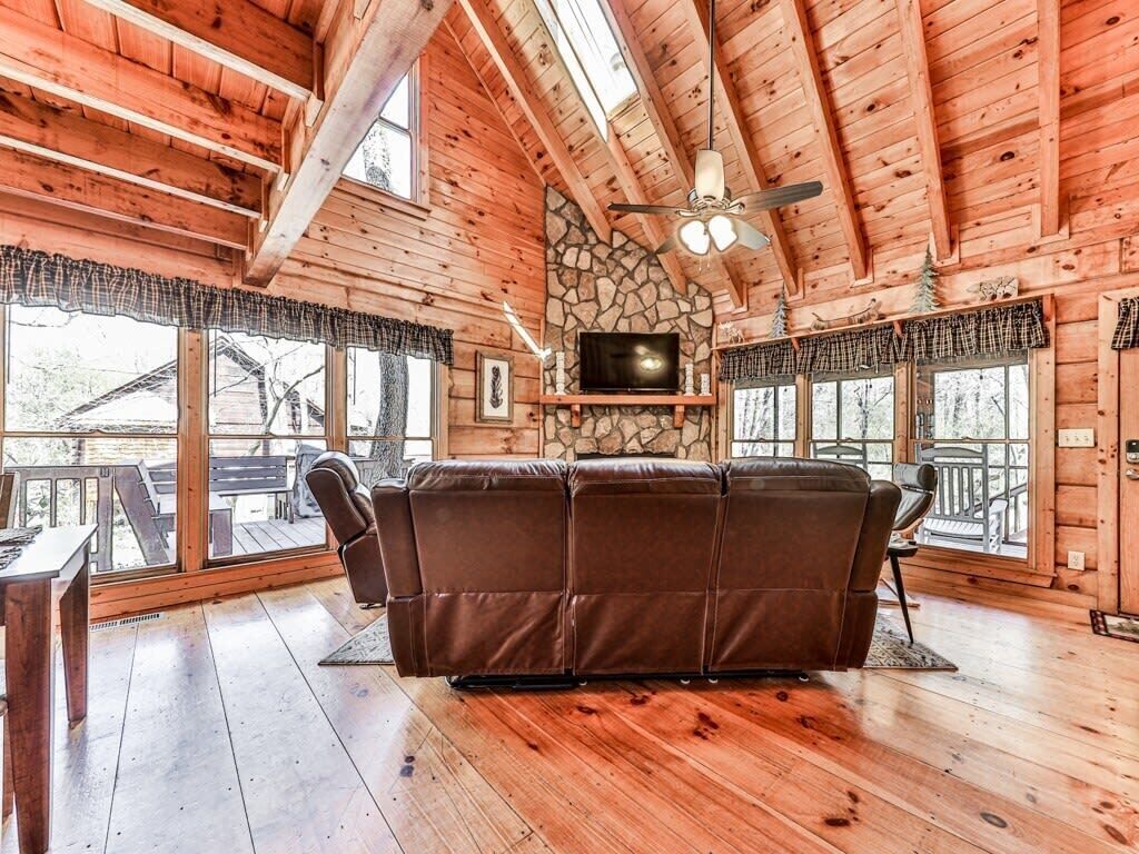 A wooden cabin interior with a stone fireplace, a brown leather sofa, and a television mounted above it.