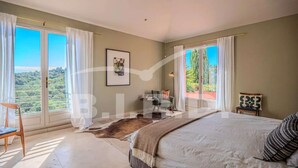 Un dormitorio con una cama grande, una silla, una mesita pequeña y vistas a zonas verdes a través de las ventanas.