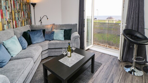A modern living room with a grey sofa, a coffee table with a bottle of wine, and a view of the sea through a large sliding glass door.