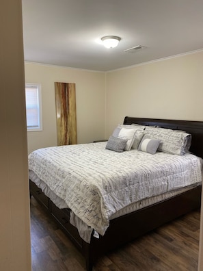Une chambre à coucher avec un lit, un plancher en bois, un luminaire au plafond, une fenêtre avec des stores et une œuvre d’art abstraite accrochée au mur.