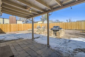 A covered patio area with a stone barbecue grill, outdoor chairs, and a wooden fence in the background. - 3bdfamily Retreat Near COS Airport & Fort Carson (Colorado Springs)