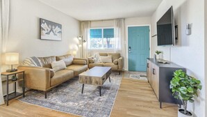 A living room with a sofa, coffee table, and TV. There is a door with a plant on top. - 3bdfamily Retreat Near COS Airport & Fort Carson (Colorado Springs)