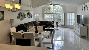 Ein modernes Wohnzimmer mit einer Eckcouch, einem Esstisch und Stühlen. Eine Kristallleuchter, ein Fernseher und ein großes Fenster mit Blick auf Grünflächen sind vorhanden.