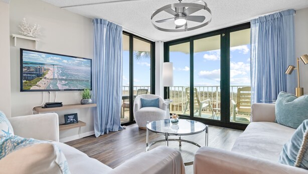 A modern living room with a large TV, comfortable seating, a glass table, and a balcony with a view. - Direct Oceanfront! Renovated! All beach gear included (North Myrtle Beach)