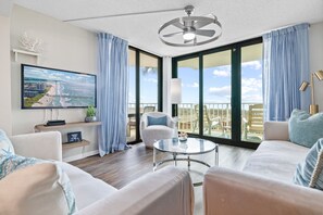 A modern living room with a large TV, comfortable seating, a glass table, and a balcony with a view.