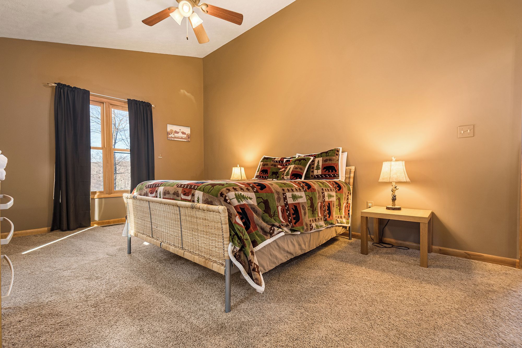 A bedroom with a wicker bed, a bedside table with a lamp, a window with curtains, and a wall-mounted ceiling fan.