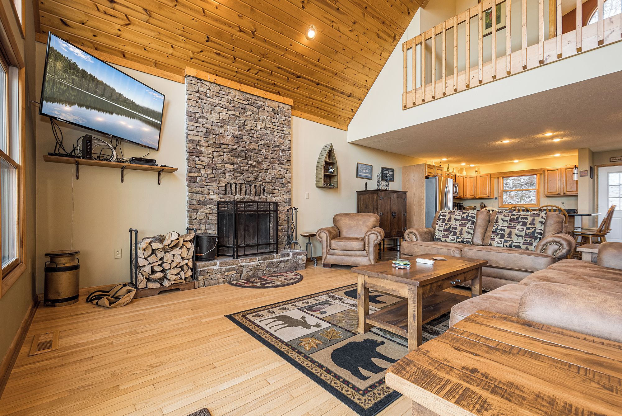 A spacious living room with a stone fireplace, wooden ceiling, and a large window overlooking a lake.