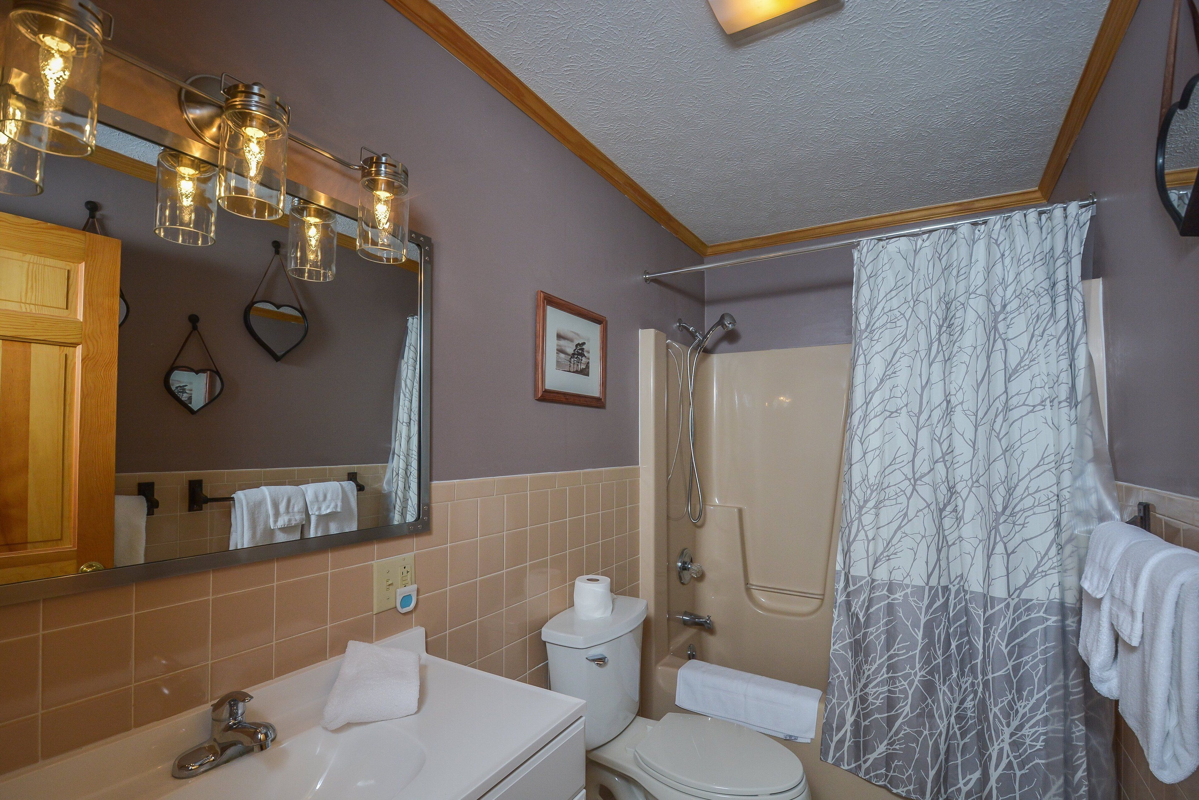 A bathroom with a sink, toilet, and shower. There are hanging lights above the sink and a shower curtain with a tree pattern.