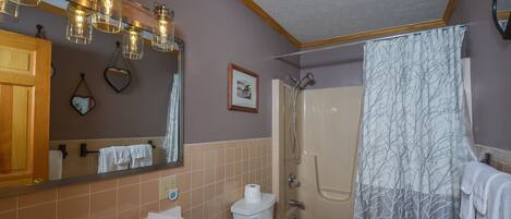 A bathroom with a sink, toilet, and shower. There are hanging lights above the sink and a shower curtain with a tree pattern.