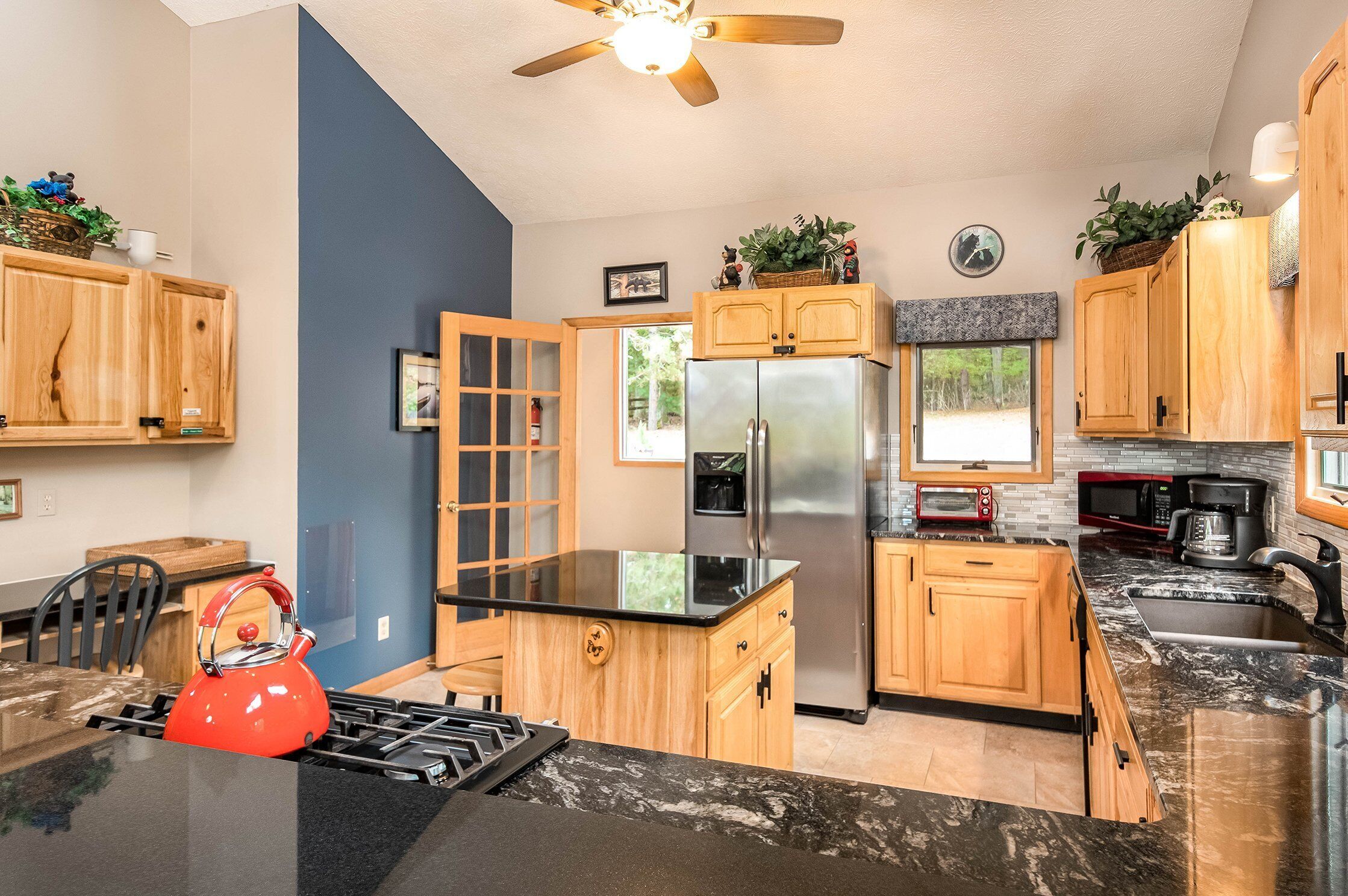 A modern kitchen with wooden cabinets, a stainless steel refrigerator, a black countertop, and a ceiling fan.