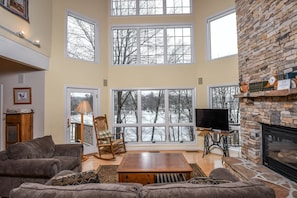Relax in a spacious living room with a high ceiling, large windows, and a stone fireplace.