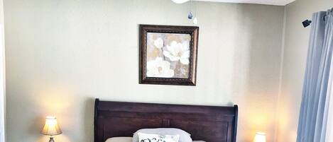 A bedroom with a wooden bed, two bedside tables with lamps, a ceiling fan, and a framed floral artwork on the wall.