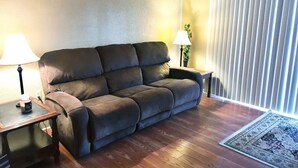 Ein Wohnzimmer mit einem dunkelbraunen Sofa, einem kleinen Holztisch, einer Stehlampe, einem Blumentopf und einem gemusterten Teppich.
