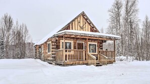 Eine Blockhütte mit Veranda und schneebedecktem Dach, umgeben von verschneiter Landschaft und kahl stehenden Bäumen.