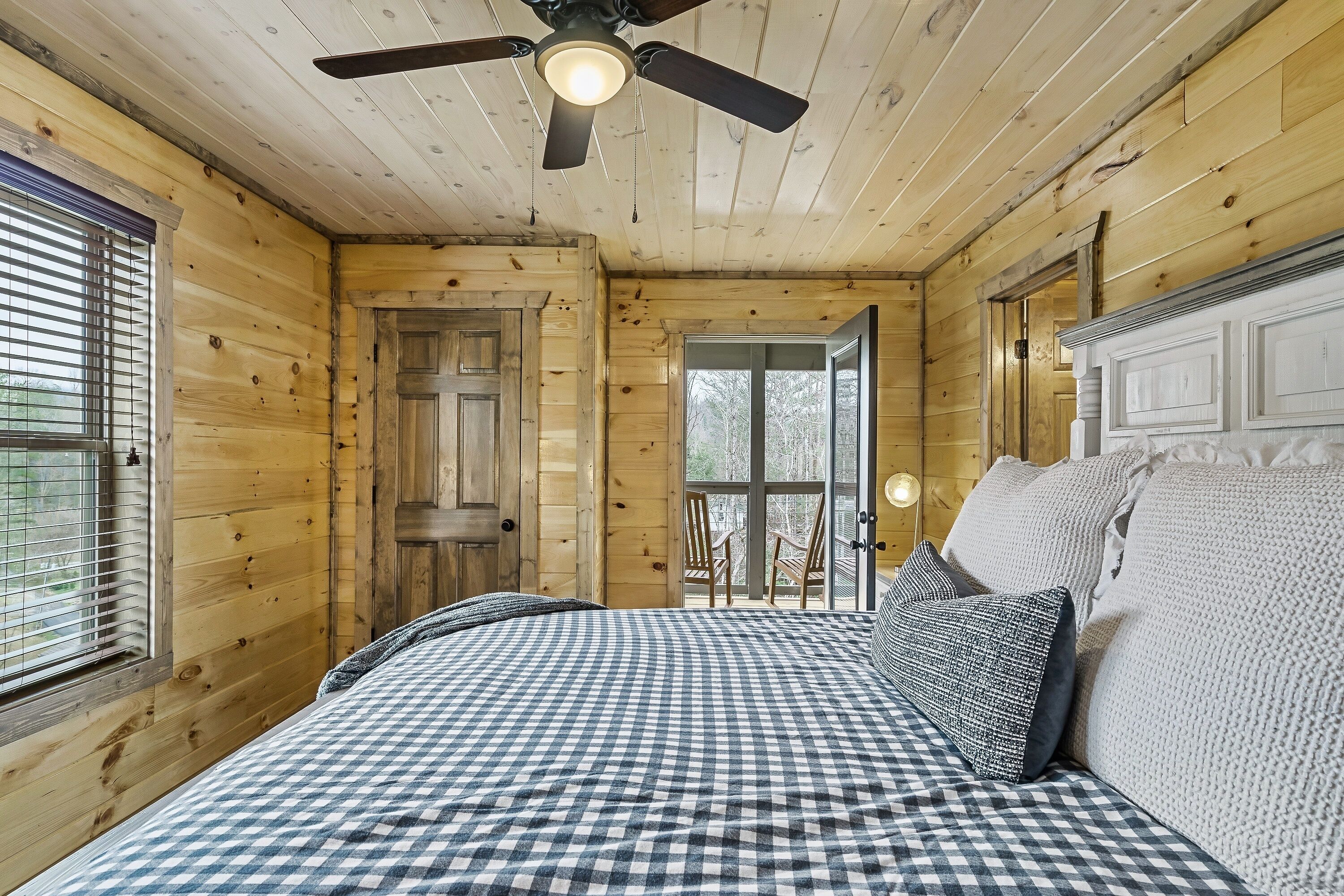 Dormitorio de una cabaña de madera con colcha a cuadros, ventilador de techo y vistas a una terraza exterior por una puerta.