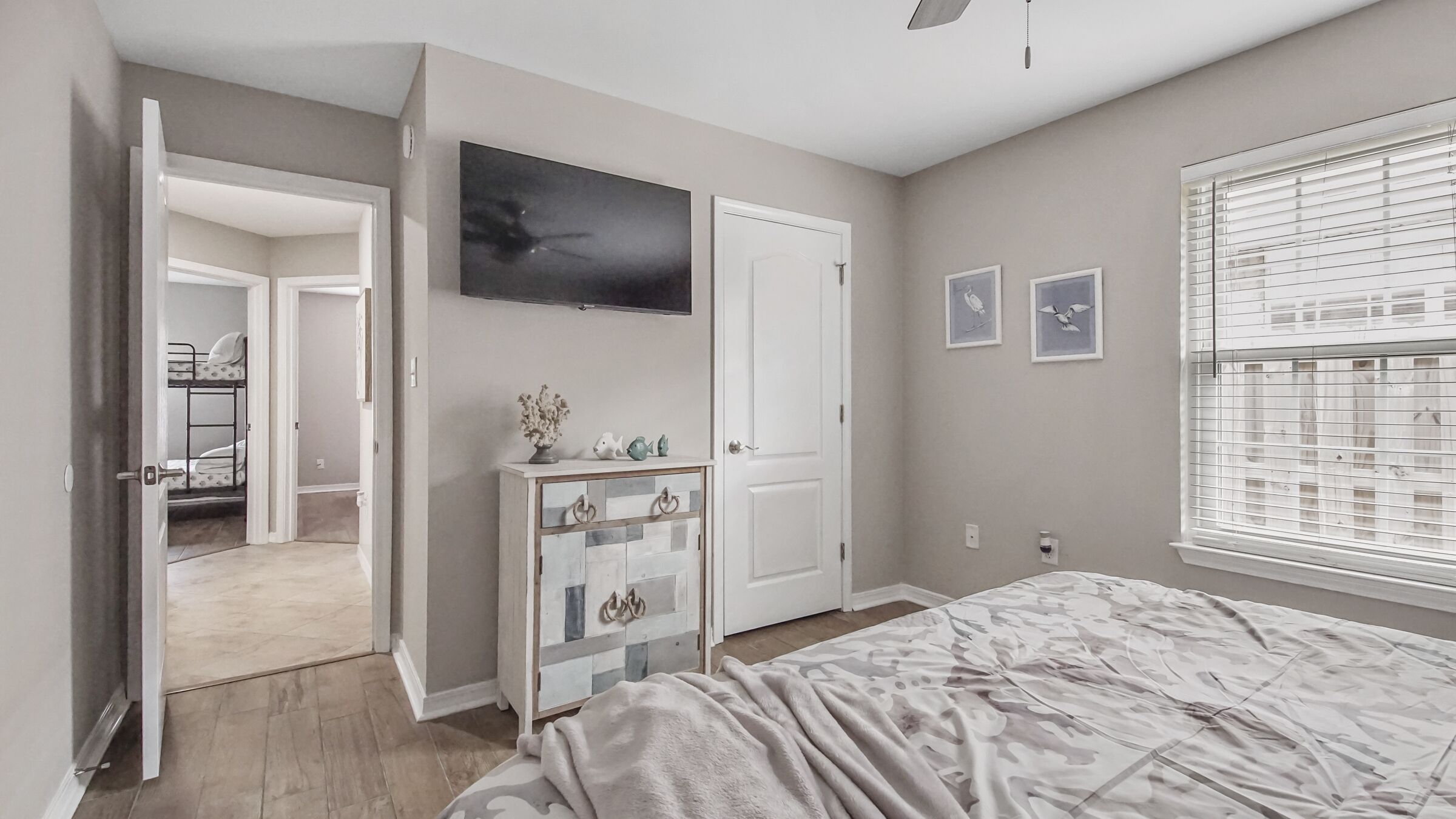 A bedroom with a bed, a dresser, a door, a window with blinds, and a hallway leading to a bathroom.
