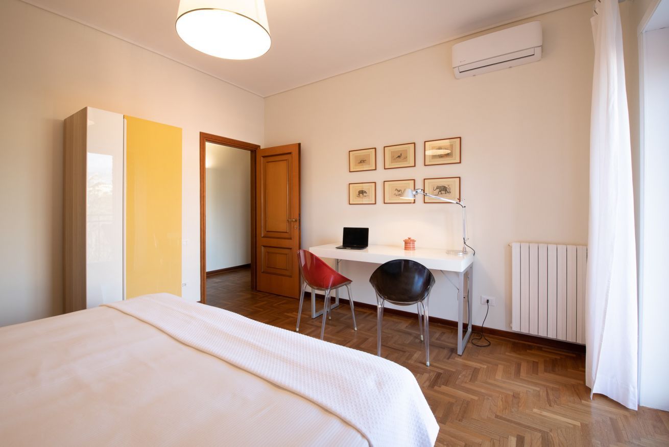 A bedroom with a bed, a desk with a chair, a radiator, and framed pictures on the wall.