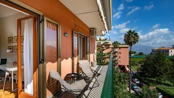 A balcony with two lounge chairs and a small table, overlooking a coastal area with palm trees and parked cars.