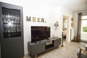 A modern living room with a flat-screen TV on a black console, a glass cabinet with wine glasses, and a 'MEER' sign on the wall.