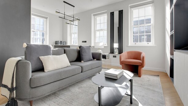Een moderne woonkamer met een grijze bank, een klein rond tafeltje en een rode fauteuil. Er zijn grote ramen met gordijnen, een zwarte radiator en een plafondlamp.